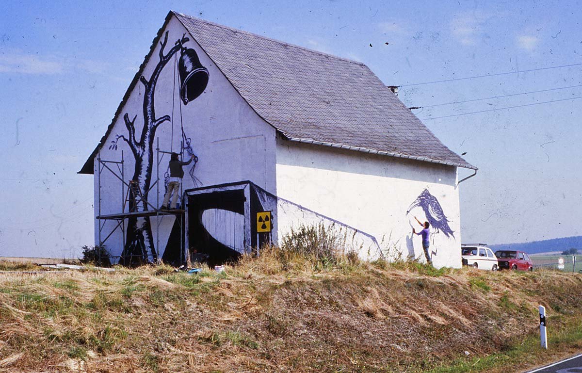 Zwei Malende an der Scheune: einer auf dem Gerüst am Giebelbild mit Baum und Glocke, ein zweiter an der Längswand; am Tor das Strahlenwarnzeichen